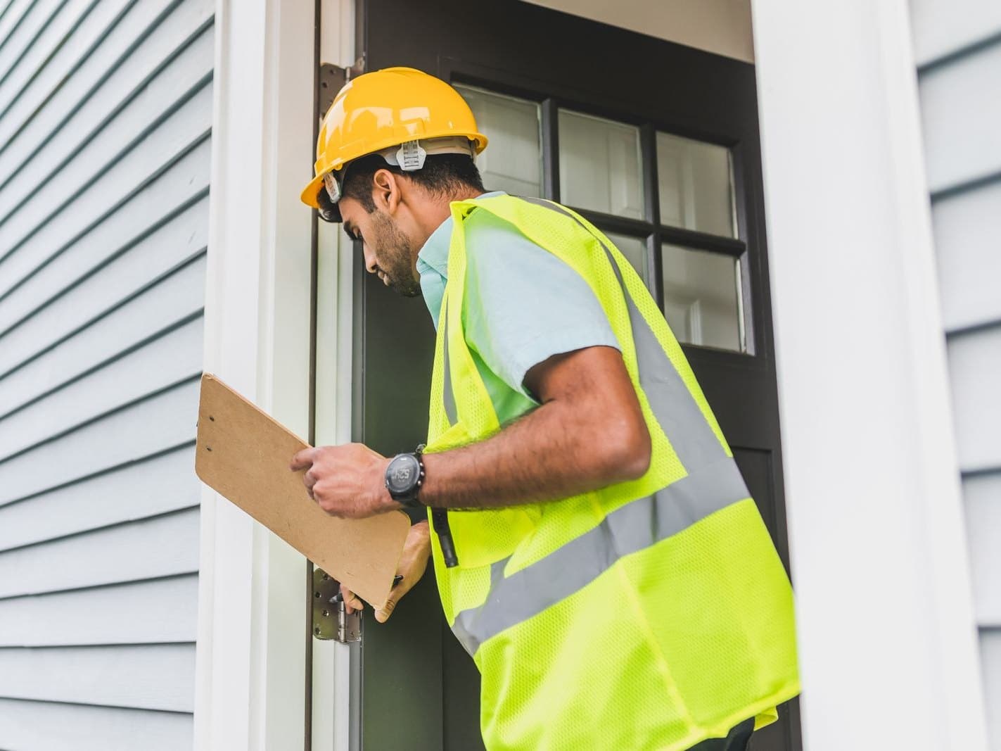 Field inspector in a hard hat and high-visibility vest documenting a property exterior on a clipboard